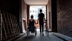 Foto: Young woman in a wheelchair holding hands with her boyfriend who walks next to her with his bike in the other hand; Copyright: Andi Weiland | Gesellschaftsbilder.de